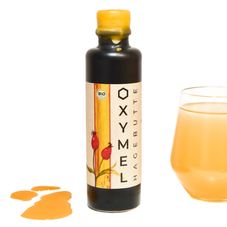 Schöne Tonflasche als Geschenk mit Bienesto Oxymel mit Hagebutte (Bio, 200ml) auf weißem Hintergrund mit einem Glas eingeschenktem Oxymel als Morgenritual im Hintergrund und ein paar Tropfen Oxymel Pur.
